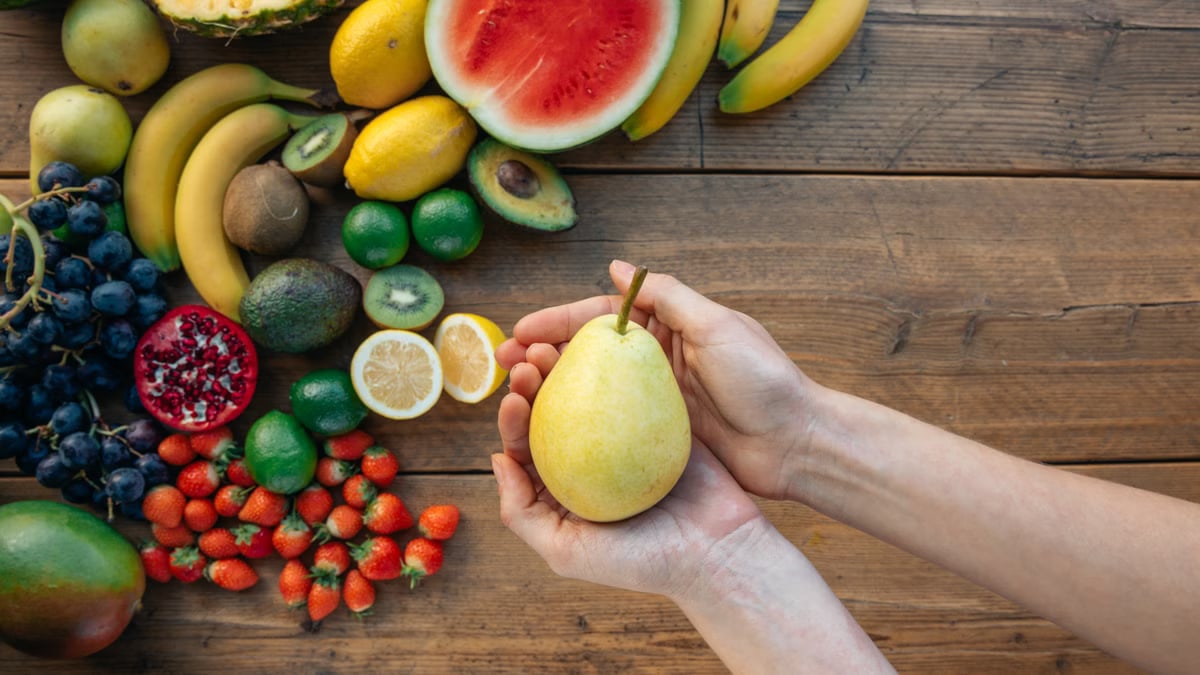 Hands holding a pear surrounded by a colorful variety of fruits on a wooden surface.